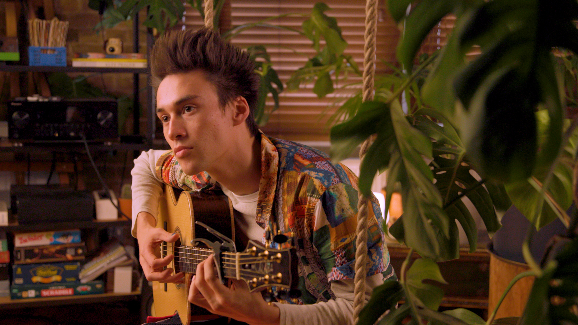 A man leans forward and strums his guitar while looking off camera. In the foreground are several green, leafy plances while a shelf of audio gear and board games can be seen in the background.