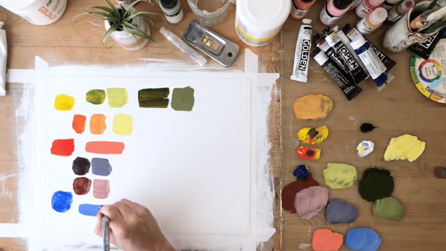 A hand adds a light blue stroke of paint onto a piece of paper taped to a wooden table. The paper is covered in different colored paint strokes. To the right of the paper is a glass paint palette with dots of different colors and above it is painting supplies like tubes of paint and a water glass.