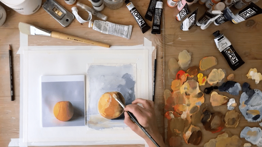 A hand holds a paintbrush, which adds beige colored paint onto a painting of an orange. To the left of the painting is a photo of an orange and to its right is mixed paint on a glass paint palette. Above the painting are various painting tools like tubes of paint and brushes.