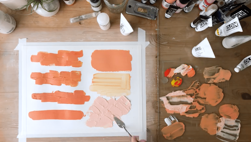 A sheet of paper is taped to a wooden table surrounded by a glass paint palette and various painting supplies like tubes of paint. A hand holding a palette knife adds thick, peach-colored paint onto the piece of paper. Strokes of orange paint of different textures and thickness fill the rest of the space on the paper.