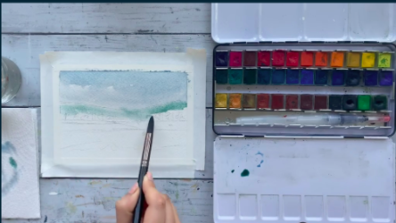 The artist uses a medium-sized brush to lay down the first layer of watercolor paint, starting with lighter colors.