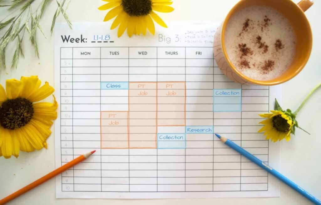 A weekly schedule laying on a white tabletop. It’s surrounded by colored pencils, sunflowers and a cup of coffee. 