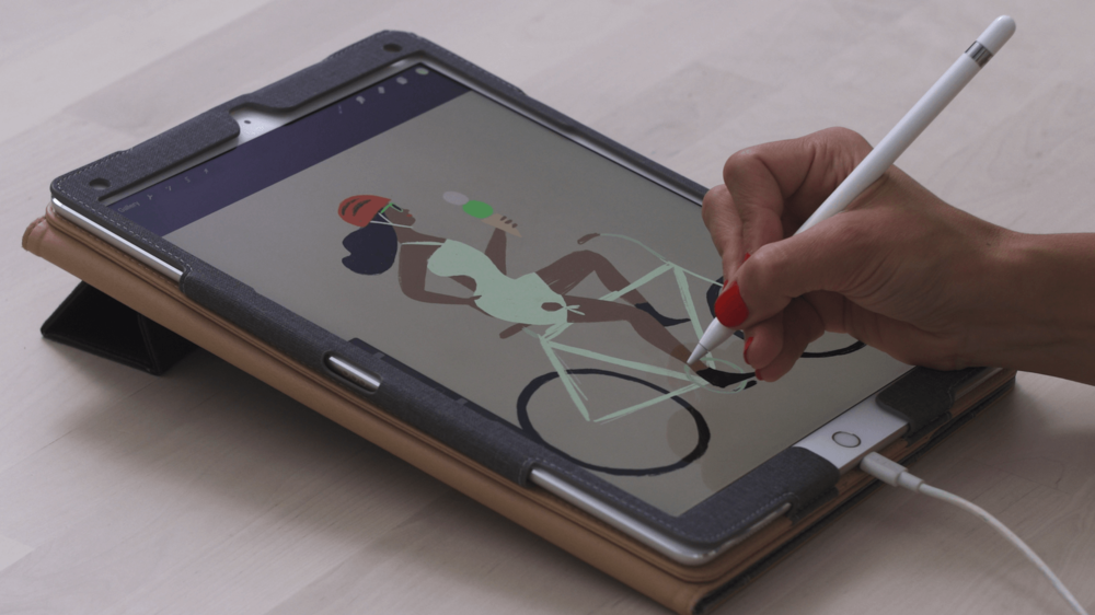 Um iPad conectado e rodando o Procreate. A mão de uma pessoa com unhas pintadas de vermelho segura uma Apple pencil sobre a tela, que exibe uma imagem de uma mulher andando de bicicleta, segurando um sorvete de casquinha.