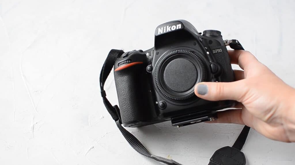 A person’s hand holding a Nikon DSLR camera body in front of a white-painted surface.