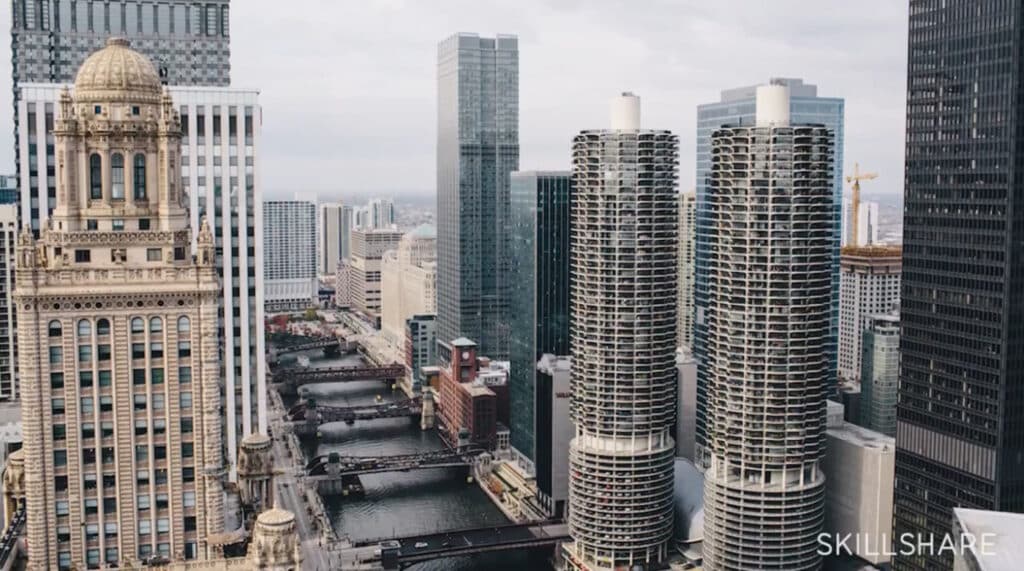 A photo of the Chicago River flanked by skyscrapers on each side, with several bridges running across it. The sky over the scene is gray and overcast.