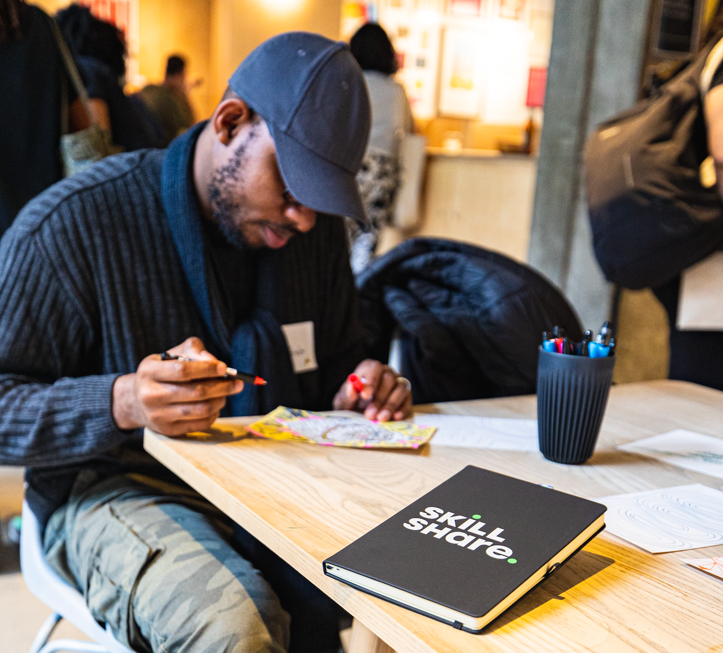 Black person with camouflage pants, navy sweater and navy ballcap, sitting at a table, looking down and doodling during the Skillshare UK Teacher Event. In front of him on the table, there's a cup with pens and a Skillshare notebook.