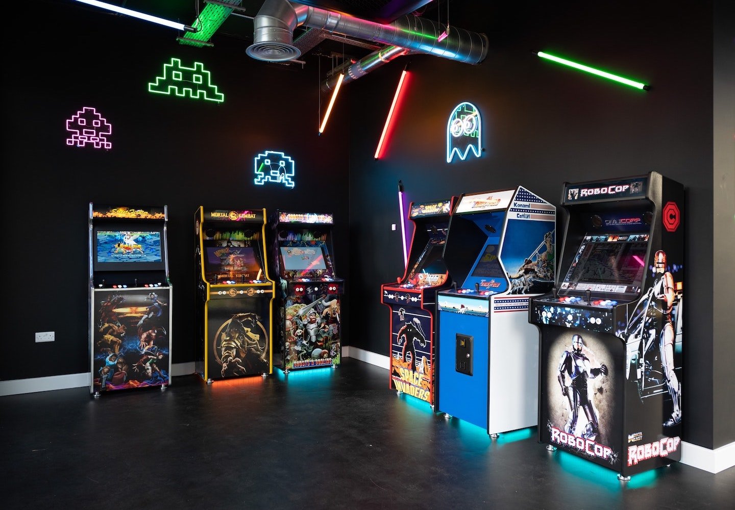 Example of a home arcade. Pac-Man and Space Invaders characters in neon lights are hung on the walls. Six cabinet video games are against two of the walls at a 90-degree angle from each other.