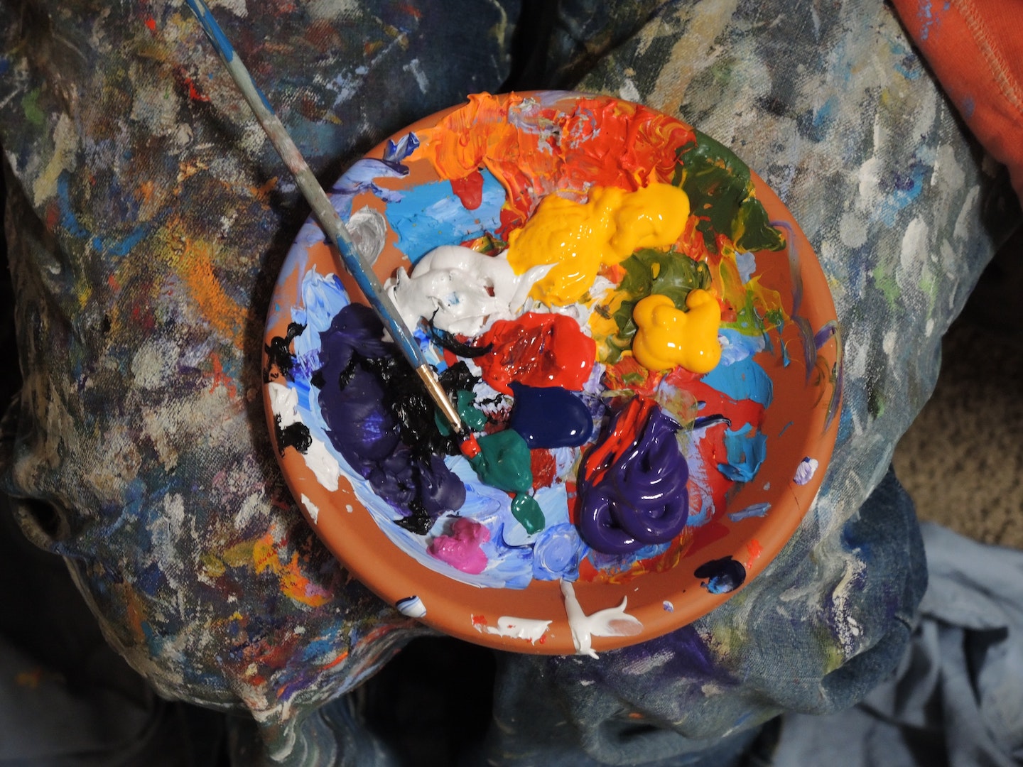 Un plato de terracota con múltiples manchas de pintura acrílica de color amarillo, rojo, azul, verde y blanco. En el borde del plato hay un pincel largo, con el pincel dentro de una mancha de pintura verde oscura. El plato está colocado sobre un paño salpicado de pintura.