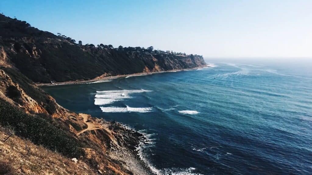 A beach landscape shot features a cliffside with trees and a deep blue body of water with gentle waves.