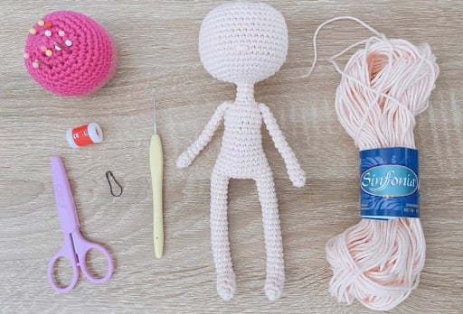 A ball of light pink yarn, scissors, a small crochet needle, a stitch counter, a pin cushion, a stitch marker, and a light pink amiguri doll body.