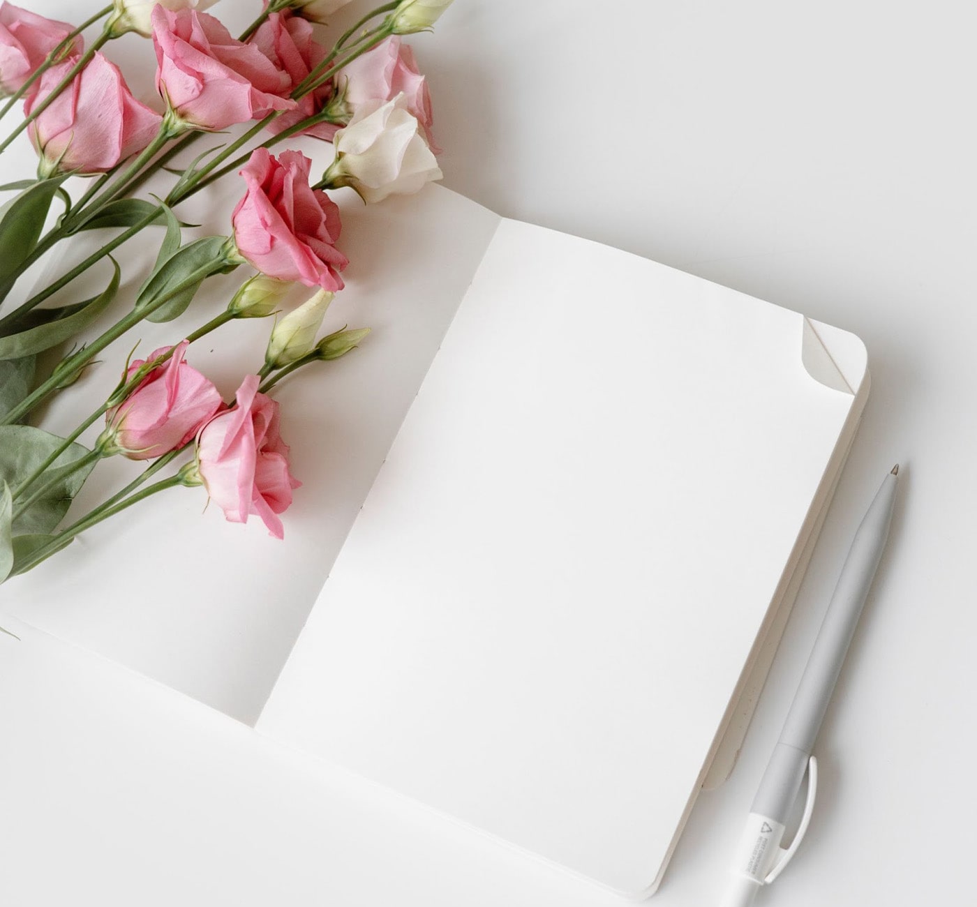 An open notebook sits on a white table with a bouquet of pink and white roses resting on its left hand page. A pen sits to the right of the notebook. 