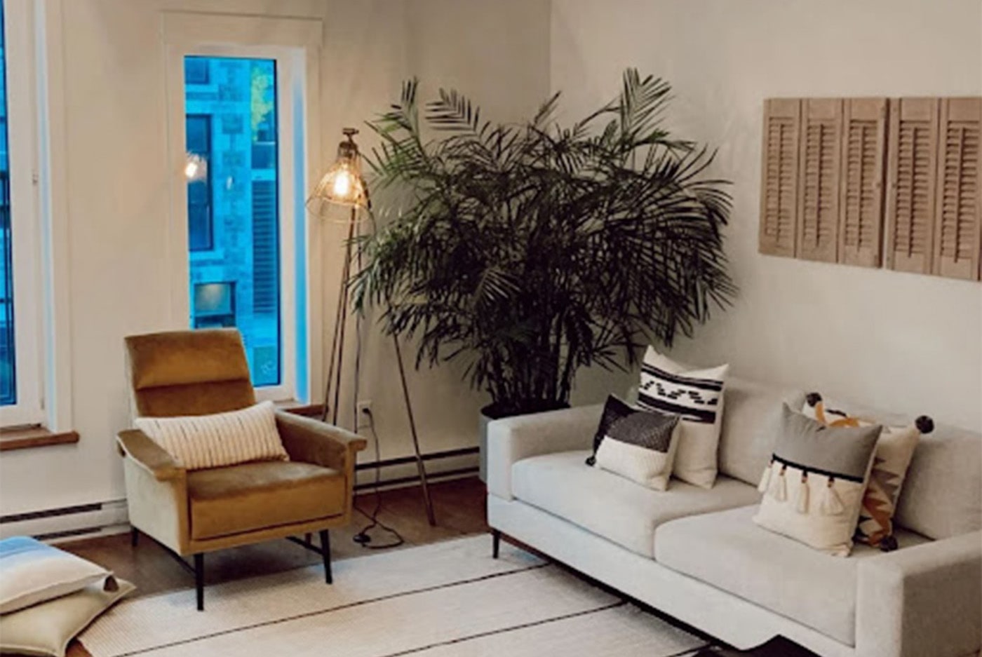 Tan and cream living room with minimalist patterns on the rug and pillows. There is a small chocolate brown velvet chair on the left wall and a cream rectangle long couch on the right wall. A large palm plant in the corner of the room with a tall floor lamp.