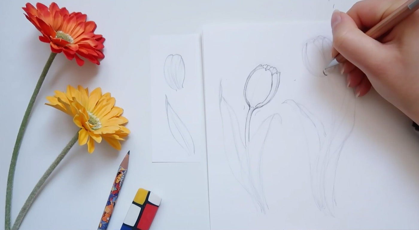 An artist draws two tulips on a piece of paper besides two faux orange and yellow flowers. 