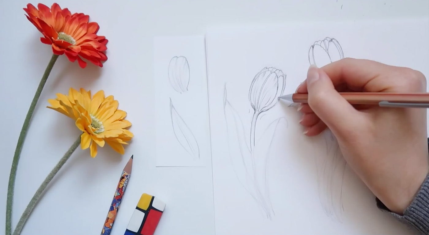 An artist adds details to a drawing of a tulip besides two faux orange and yellow flowers. 