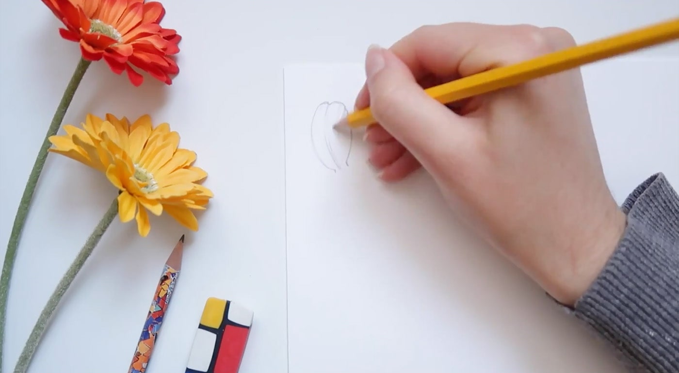 An artist fills in a tulip petal with a few curved lines on a piece of paper sitting beside two faux orange and yellow flowers. 