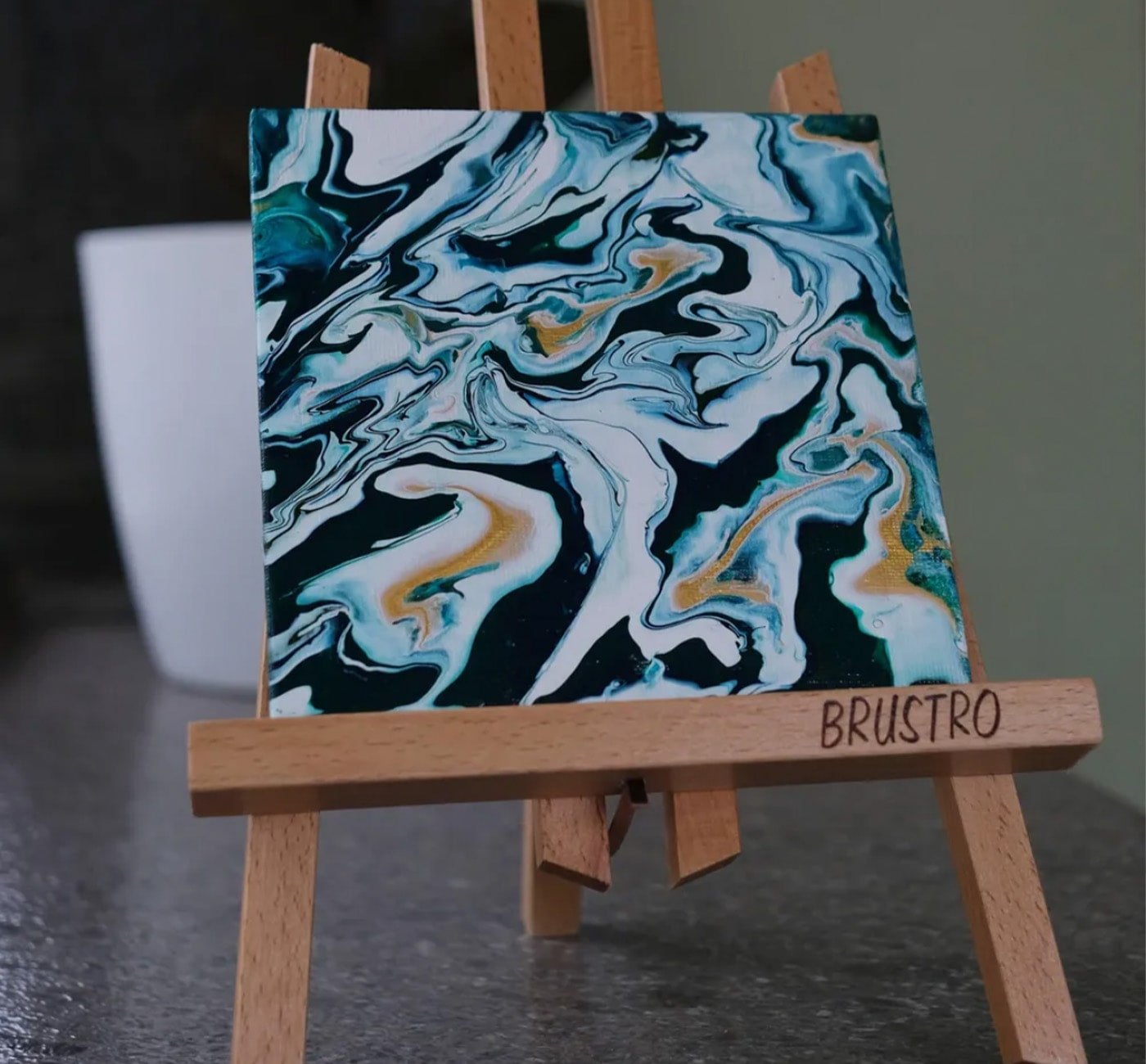 Finished fluid painting in black, white, gold , and teal paint mounted on a small easel.  Fluid painting looks like marble it's just a bunch of swirls of painting covering the whole painting fluidly.