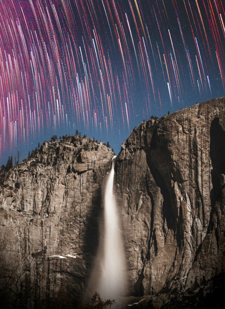 Two large cliffs sit with a waterfall in between them. Colorful star trails cover the sky with reds, purples, oranges and whites.