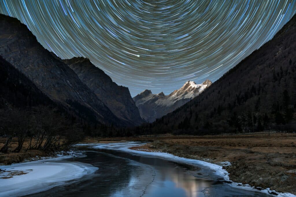 An icy river flows through a valley surrounded by snow-capped mountains. The sky is covered in blue, green and white star trails that turn in a circular motion. 