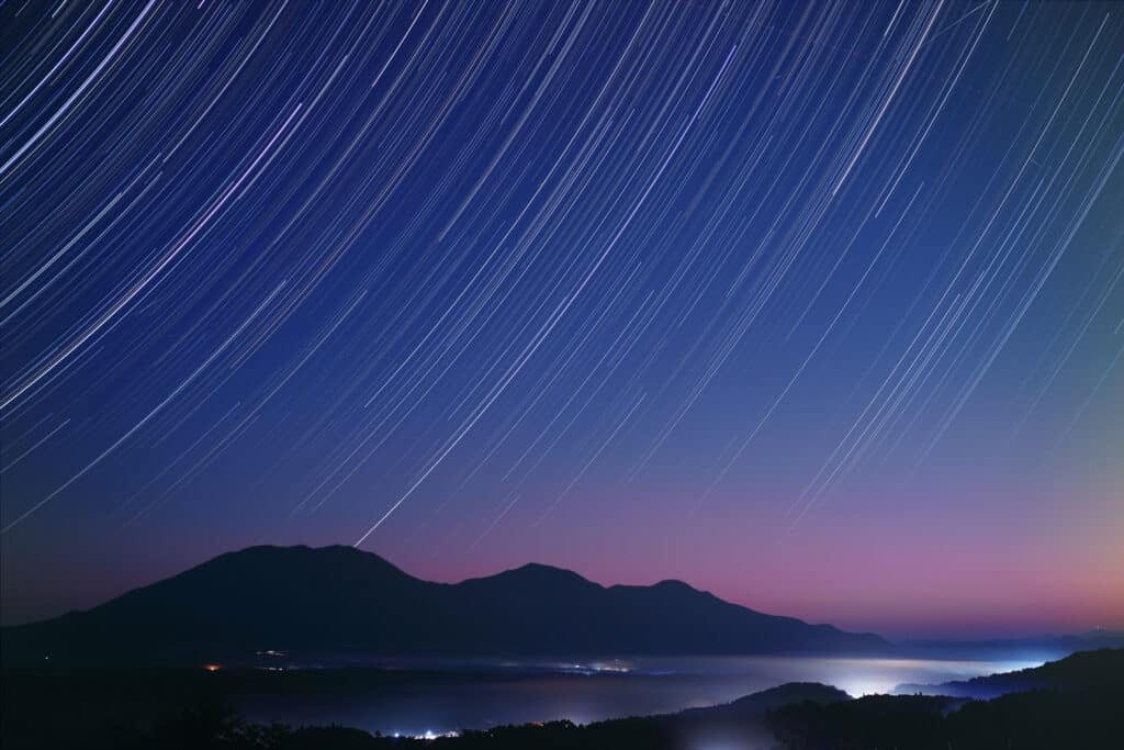 Blue and pink night sky full of curved star trails over a landscape of mountains and a lake