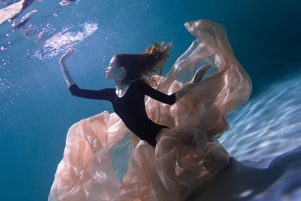 A model in a black bodysuit poses underwater, surrounded by a peach-colored fabric.