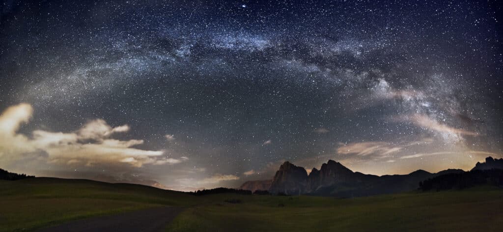 Dark, partly cloudy night sky with the Milky Way arching over a landscape of mountains and grassy hills