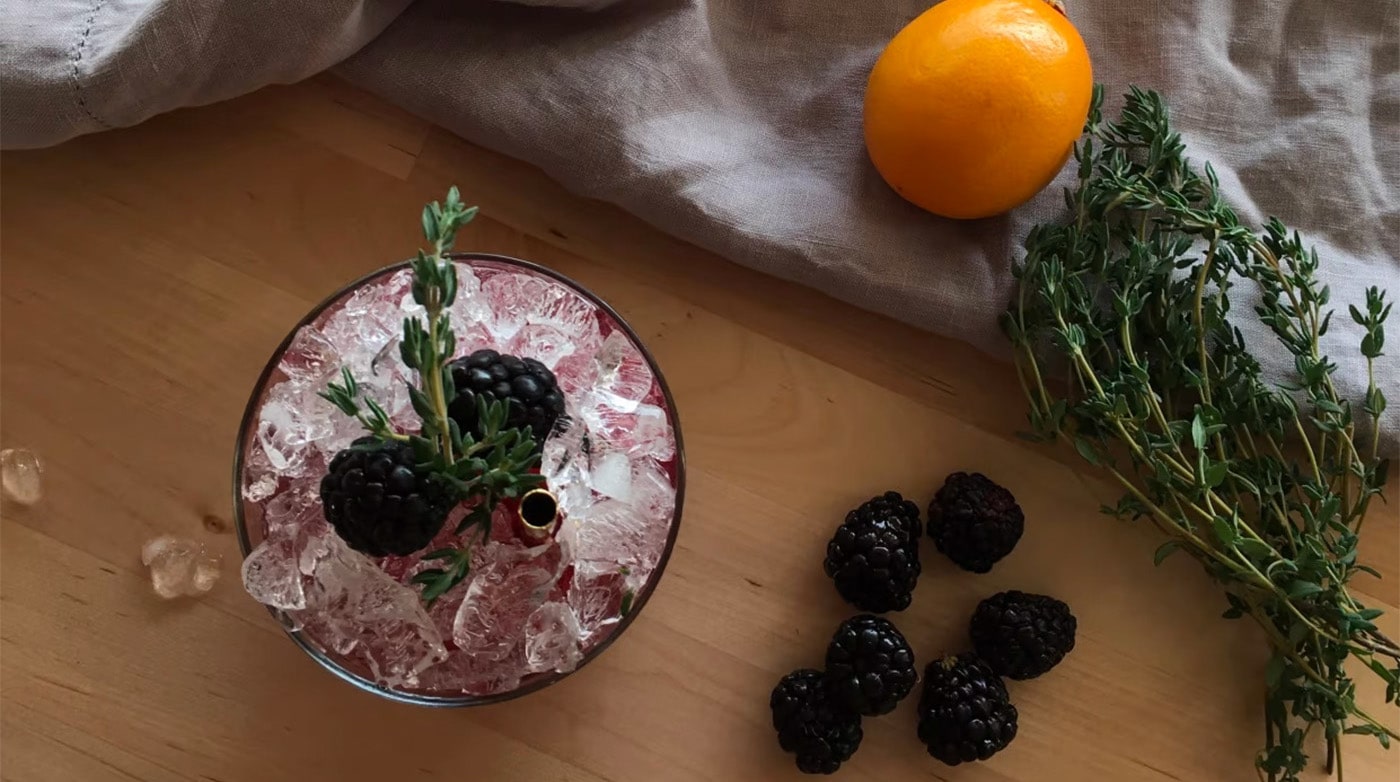 Blackberries, rosemary, and an orange sit on a table around a fancy alcoholic drink with blackberries on top.