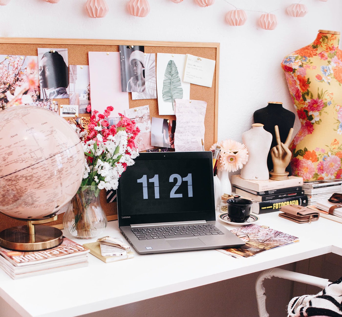 A desk with lots of things on it but most are pink. In the center of the desk is an open laptop with a flip clock showing the time on it. Behind that is a bulletin board of images. On the left of the laptop is a vase of flowers and a pink globe. To the right is a coffee cup, a wallet, and some jewelry busts and books.