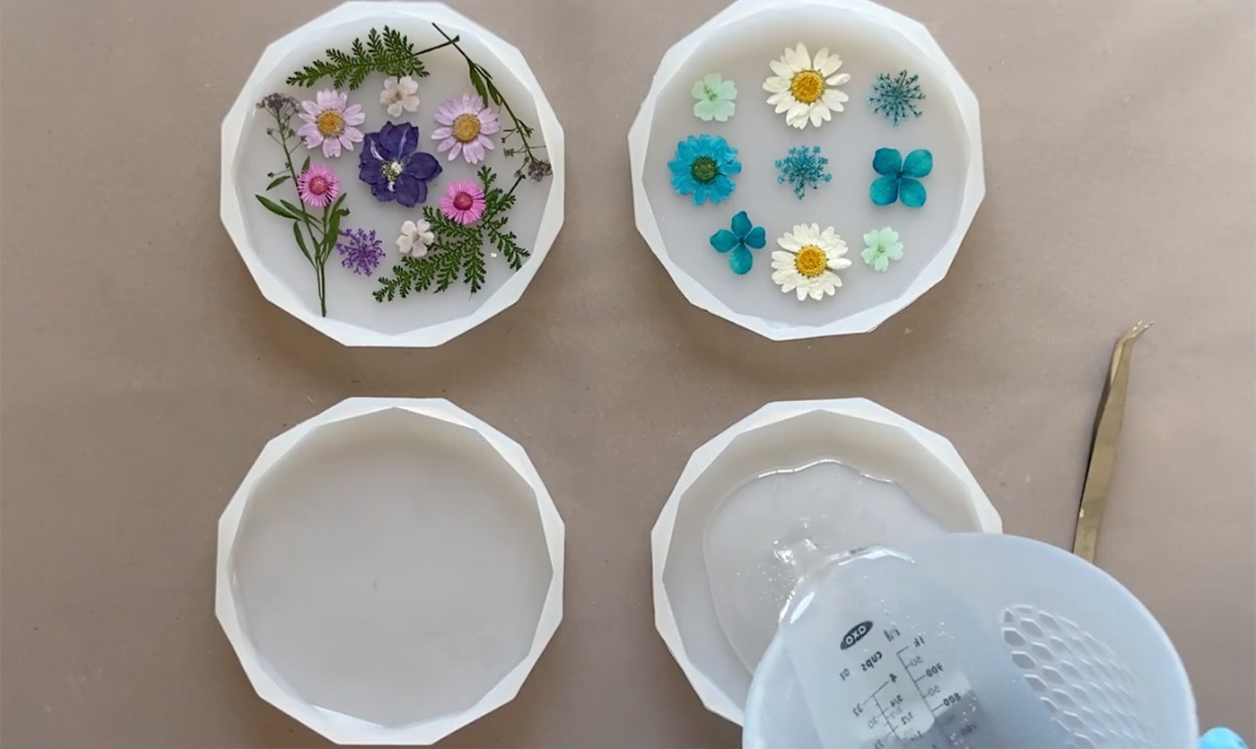 Geometric molds with dried flowers placed in them. Person is pouring resin over them slowly but not filling all the way. Tweezers are on the side