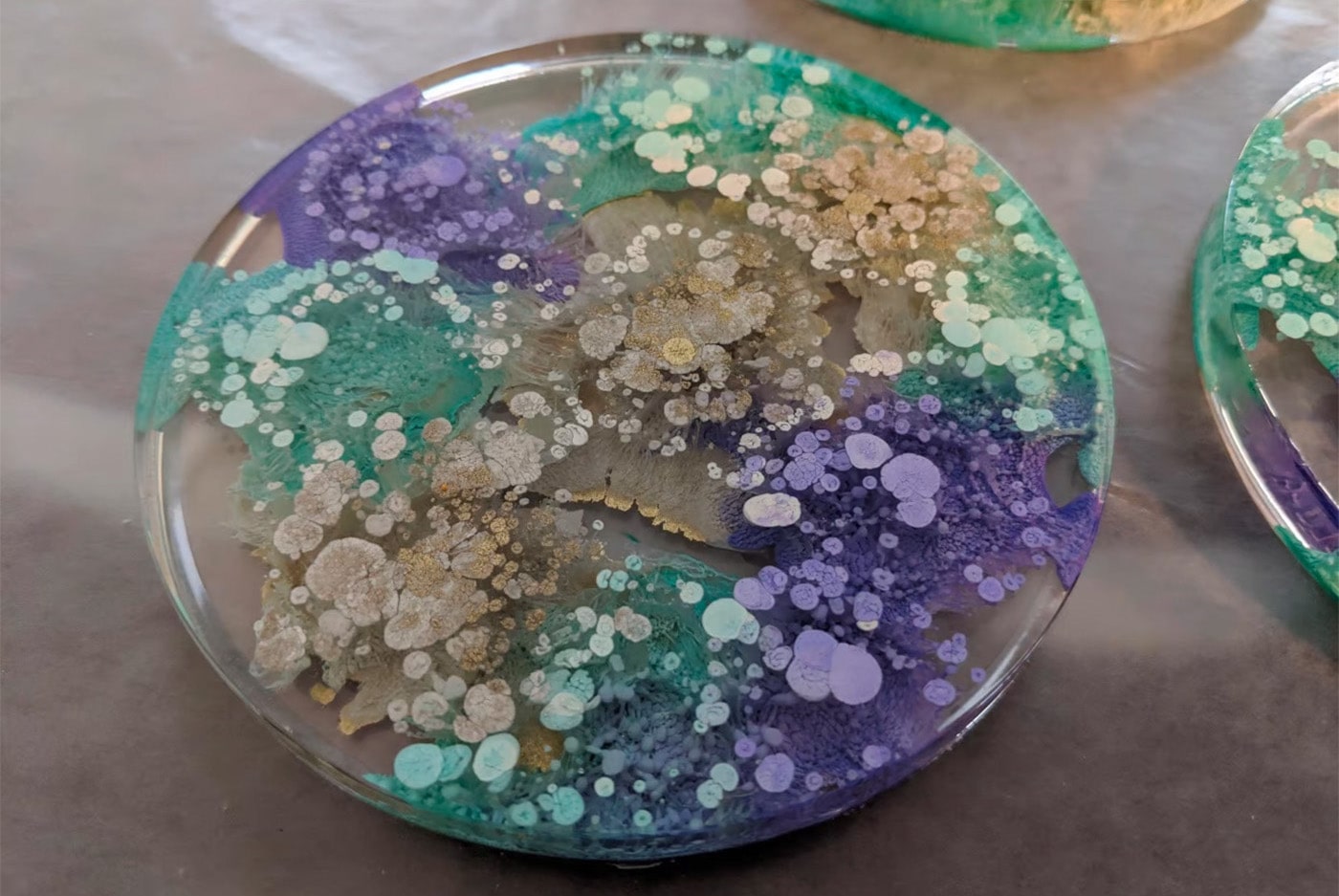 Resin coaster with paint blobs in it. Paint is purple, gold, and green.