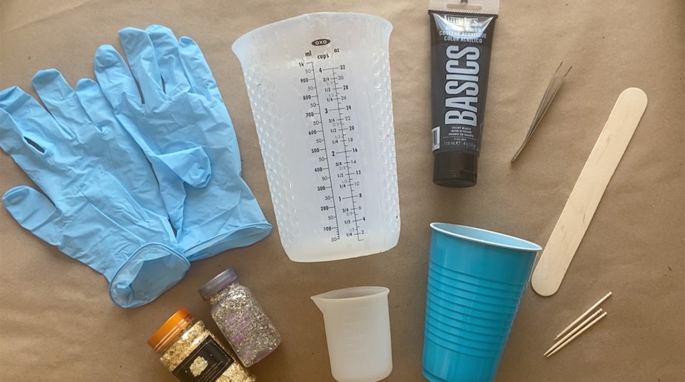 Materials for resin include: gloves, measuring cup, cup, paint, popsicle stick, glitter, toothpick, tweezer, smaller measuring cup.
