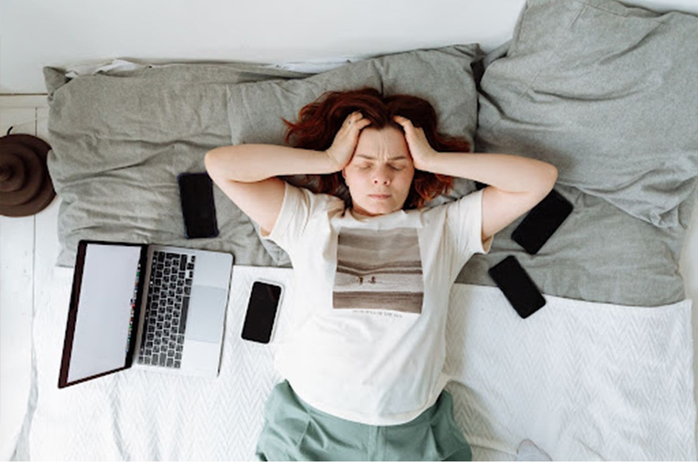 Girl laying on a bed with a distressed look on her face. Eyes closed, both hands pushing her hair back. Open Laptop and 4 cell phones surrounding her.