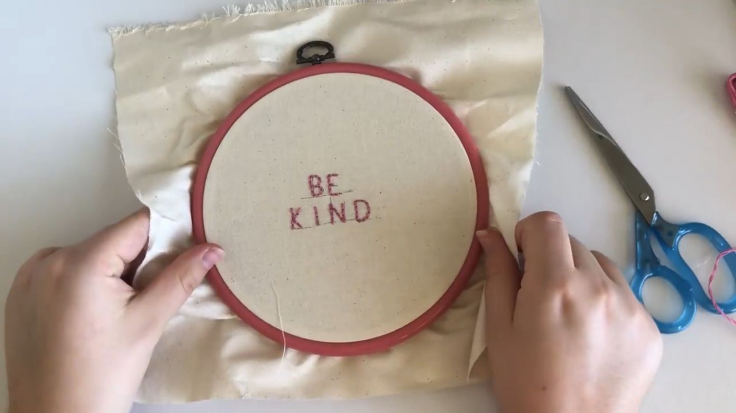 Hands holding an embroidery hoop, with the words ‘be kind’ embroidered in all-capital block letters on the fabric in pink thread.