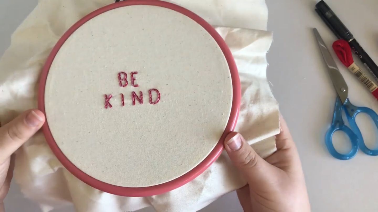 Hands holding an embroidery hoop, with the words ‘be kind’ embroidered in all-capital block letters on the fabric in both pink and red thread.