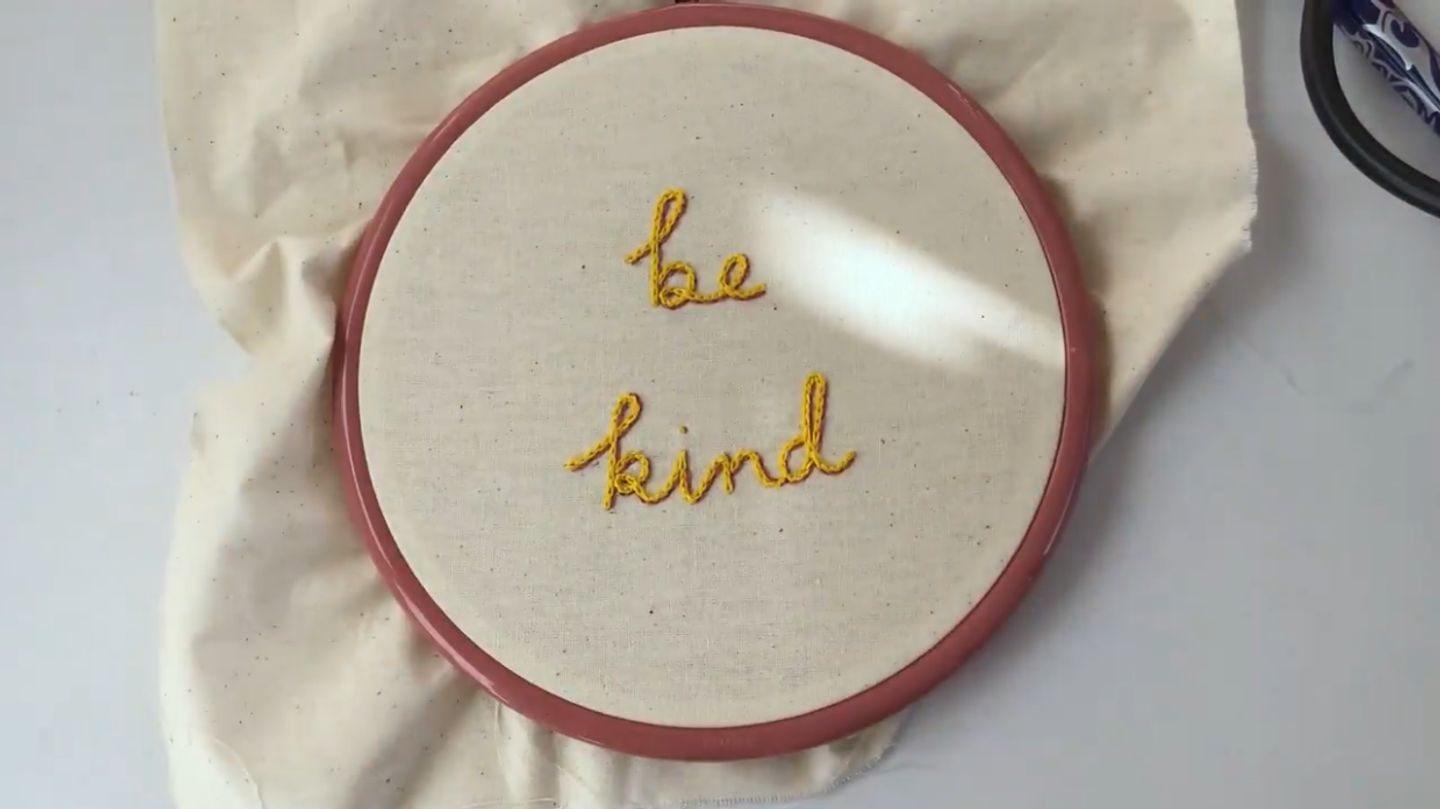 An embroidery hoop on a white background, with the words ‘be kind’ embroidered on the fabric in both yellow and pink thread for a 3D effect.