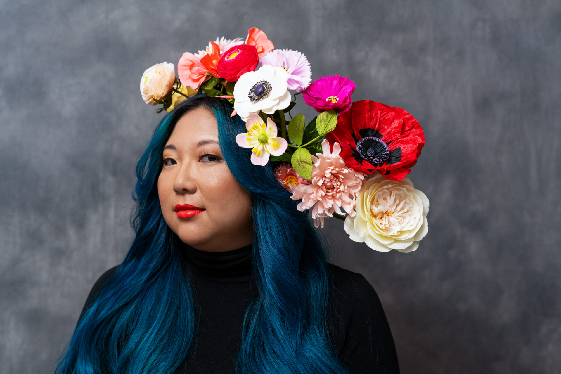 A woman with long vibrant blue hair models a head piece made of many colorful paper flowers.