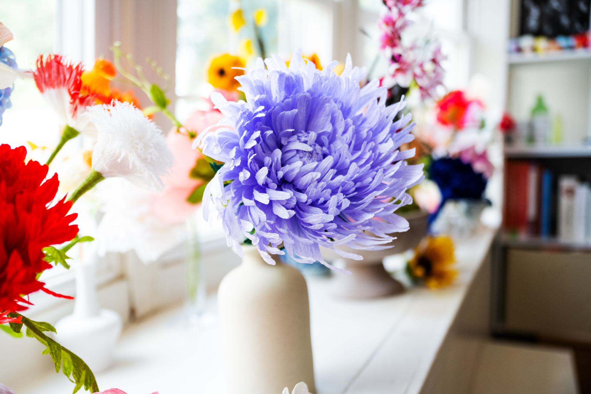 An assortment of colorful paper flowers in white vase in vases on a shelf.