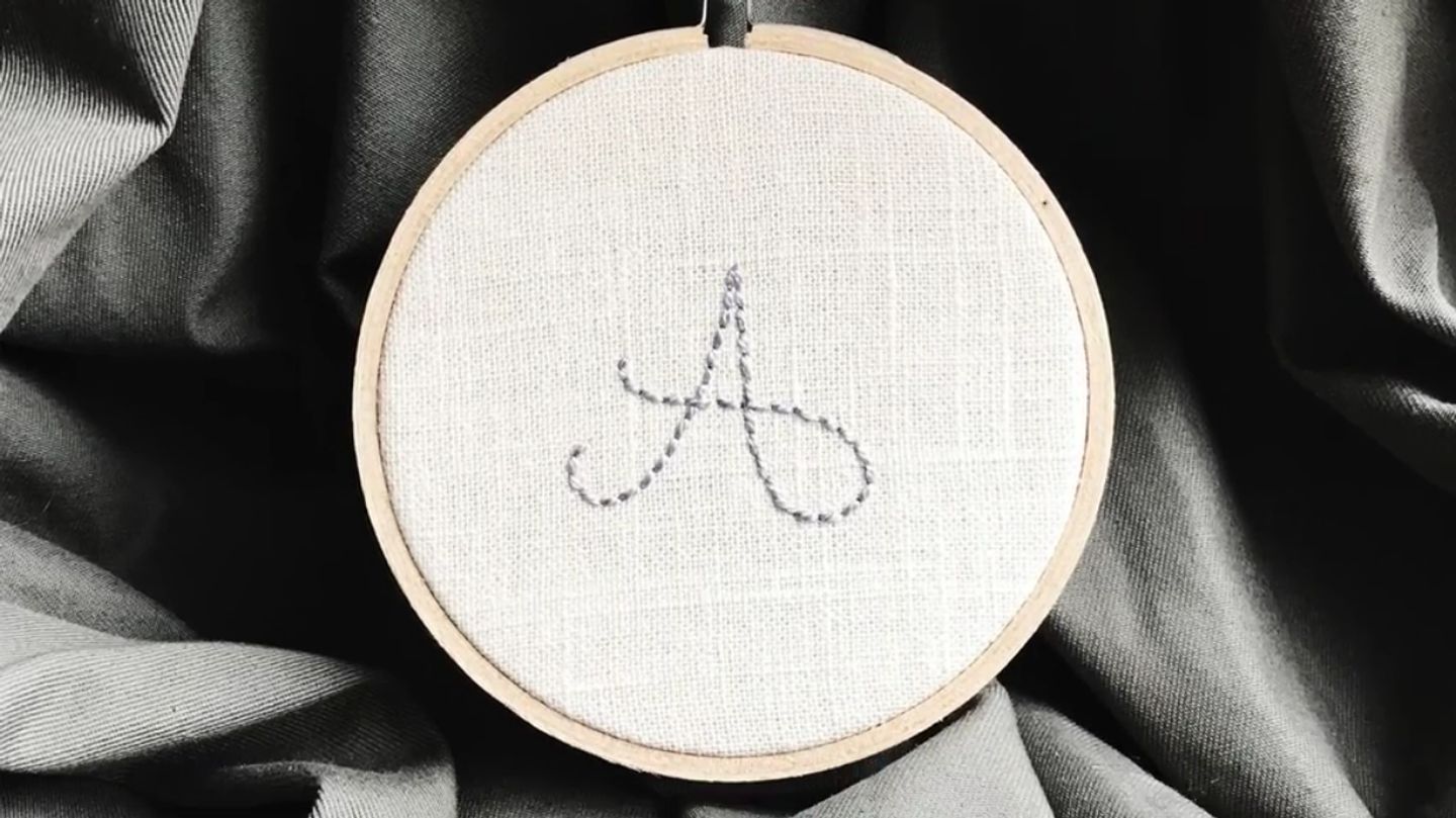 An embroidery hoop on a gray cloth background, with a cursive letter ‘A’ stitched onto the fabric in gray thread.