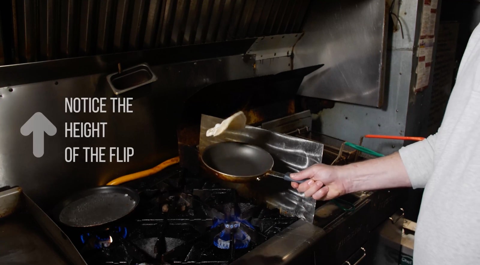 Someone flips a fried egg in a pan in a commercial kitchen. An arrow and the words “Notice the height of the flip” are written on the left side.