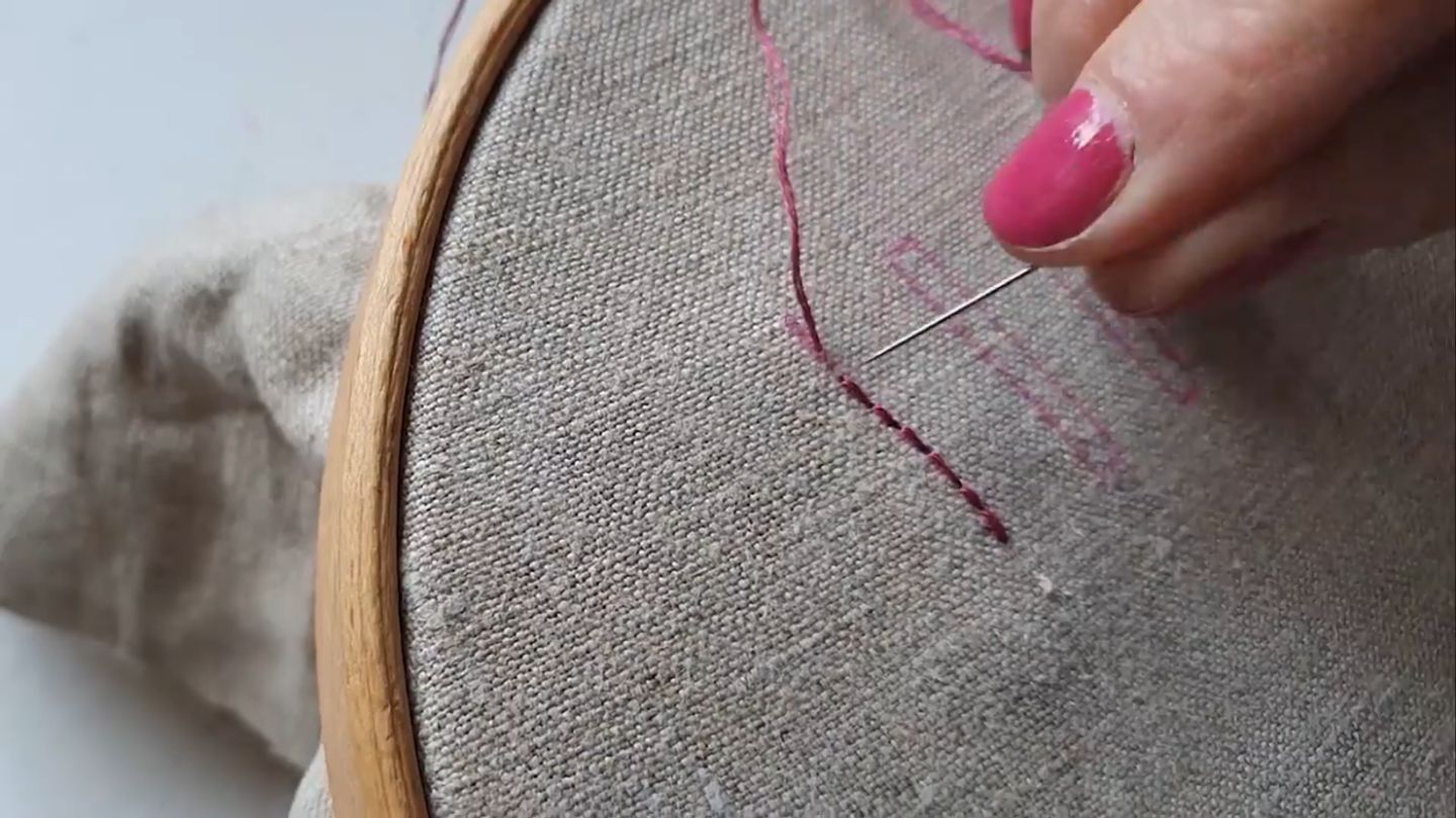 A woman’s hand using a needle and pink embroidery floss to stitch a solid line on a piece of natural-colored fabric using the back stitch. 