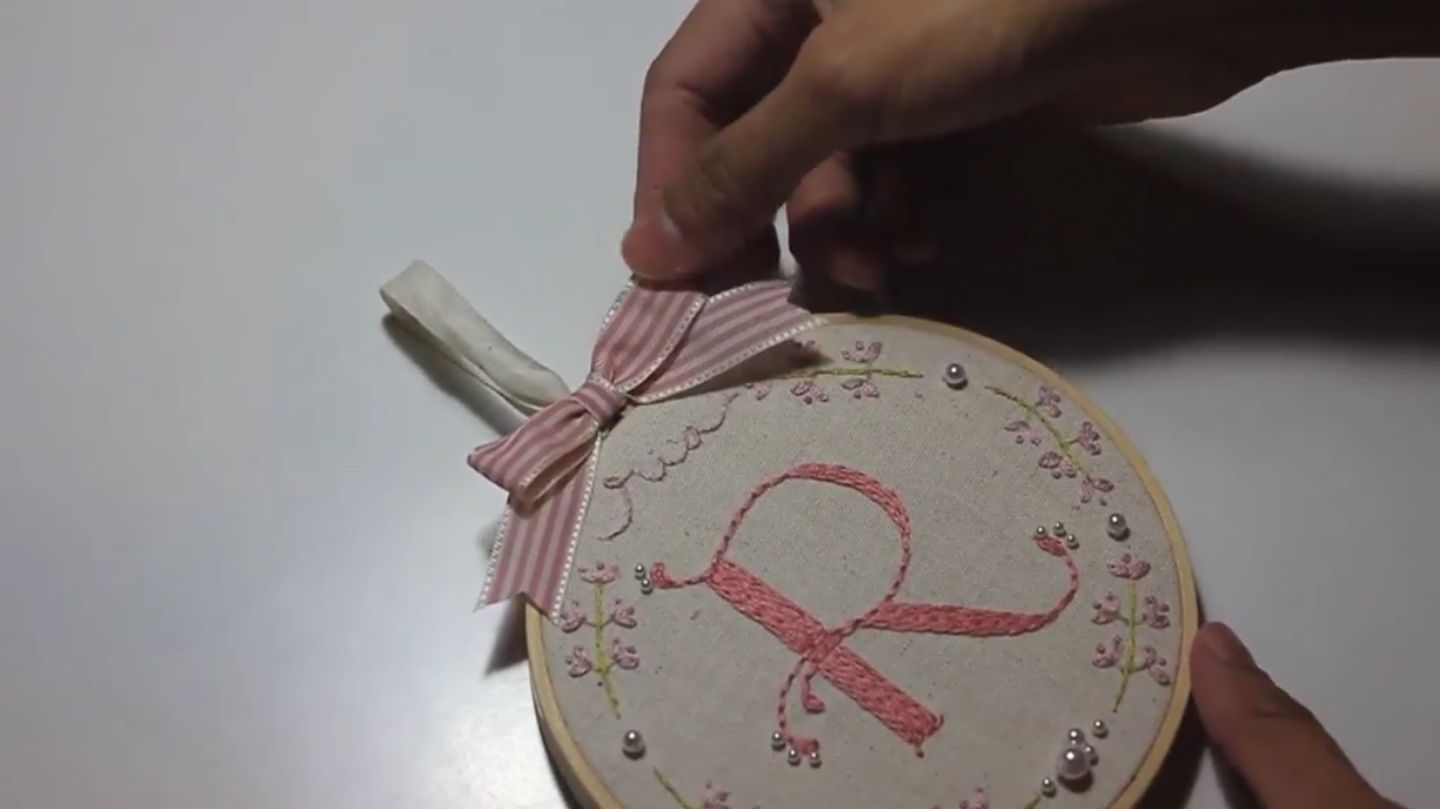 Hands hold an embroidered letter "R" in pink on a cloth in an embroidery hoop with a pink bow at the top.