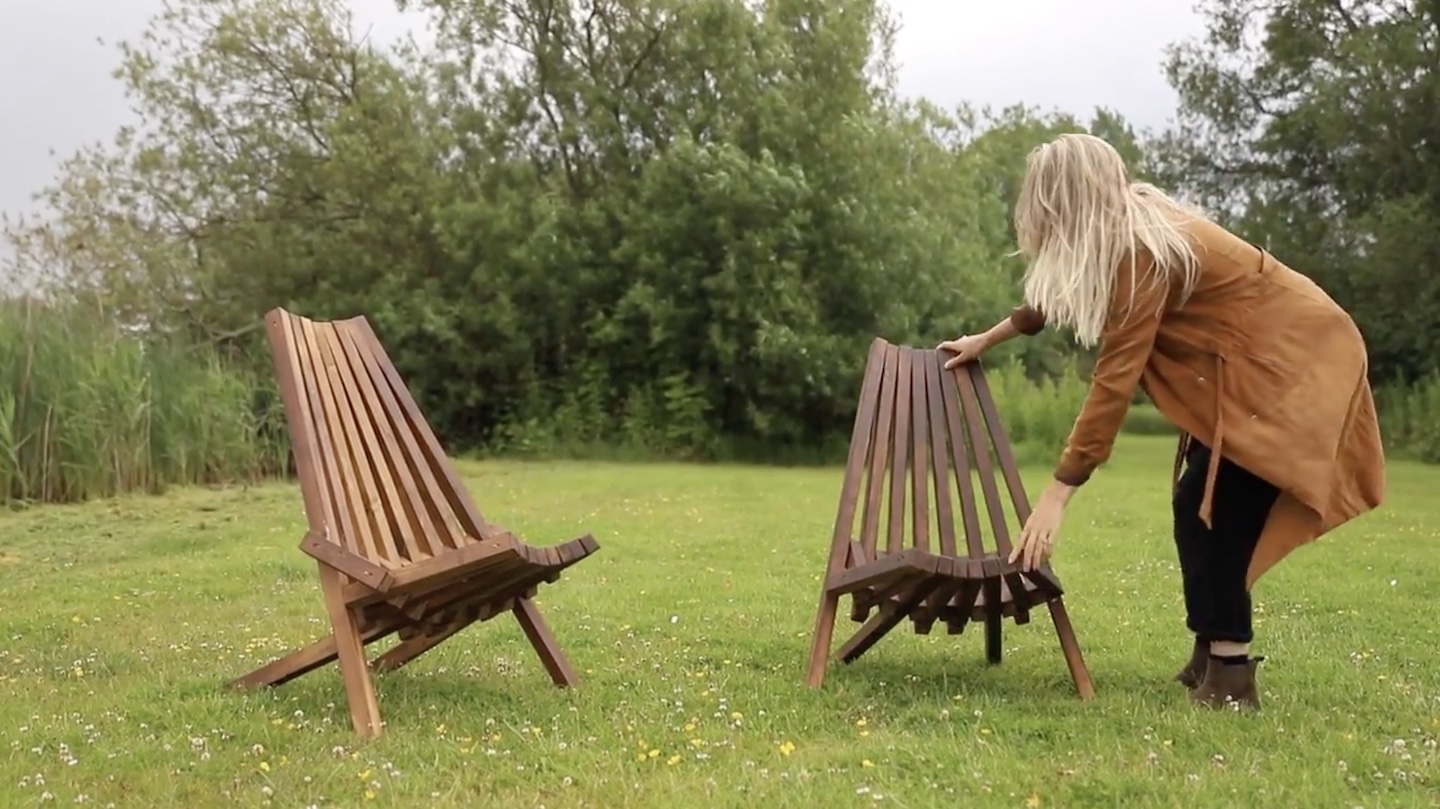 A wooden chair sits in a lush green garden. A woman with blonde hair and a rust-colored jacket sets a second wooden chair next to the first one.
