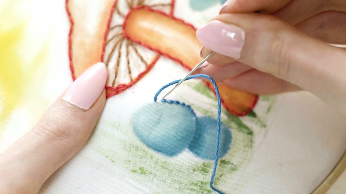 A pink-nailed hand outlines a light blue hand-painted mushroom with stitches of bright blue thread.