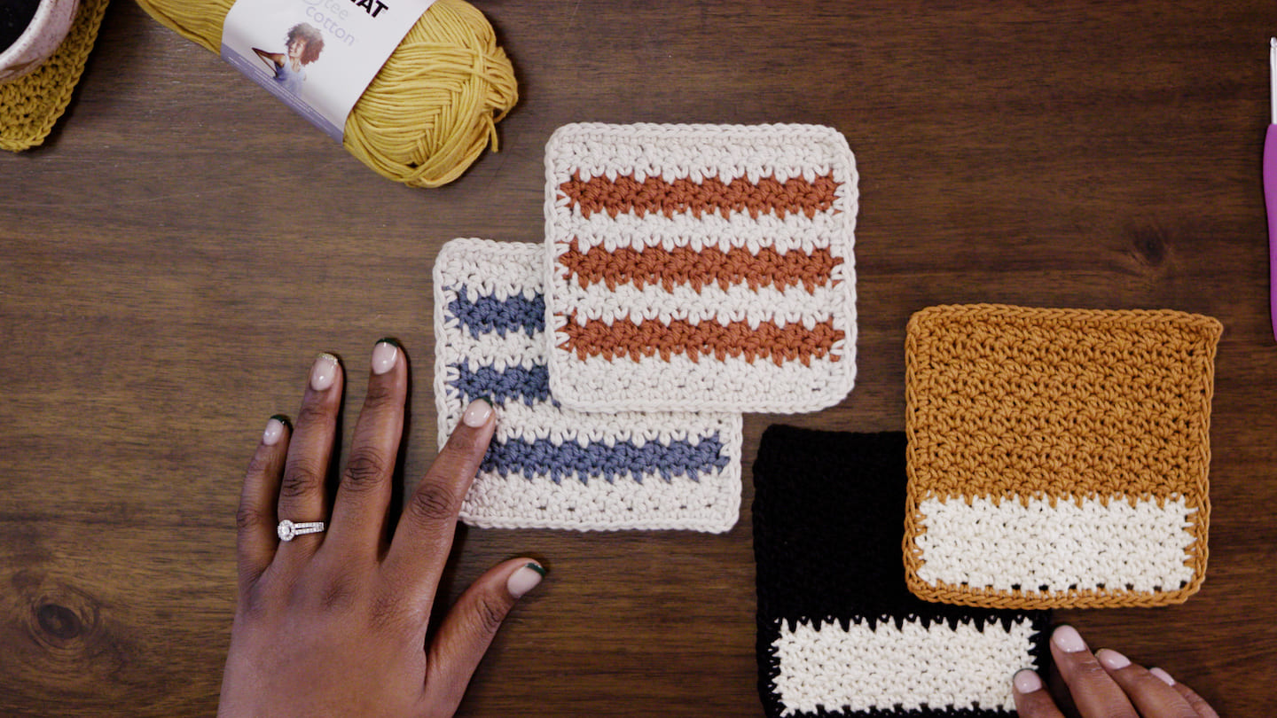 Four square-shaped crochet coasters sit on a wooden table next to a bundle of yellow yarn and two hands ready to get crafting. The first two coasters are striped white and blue and white and red. The last two are color-blocked. One is black and white and the other is yellow and white.