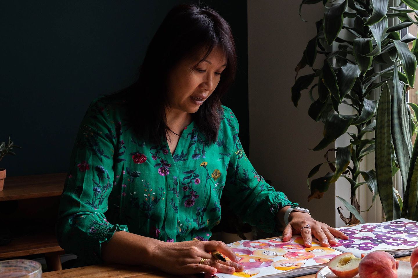 Ohn Mar Win sitting at a table with a sketch book open in front of her. On the table is a plate of peaches, some whole, some cut in half. The sketch book page contains illustrations of the peaches in shades of yellow, pink and purple.