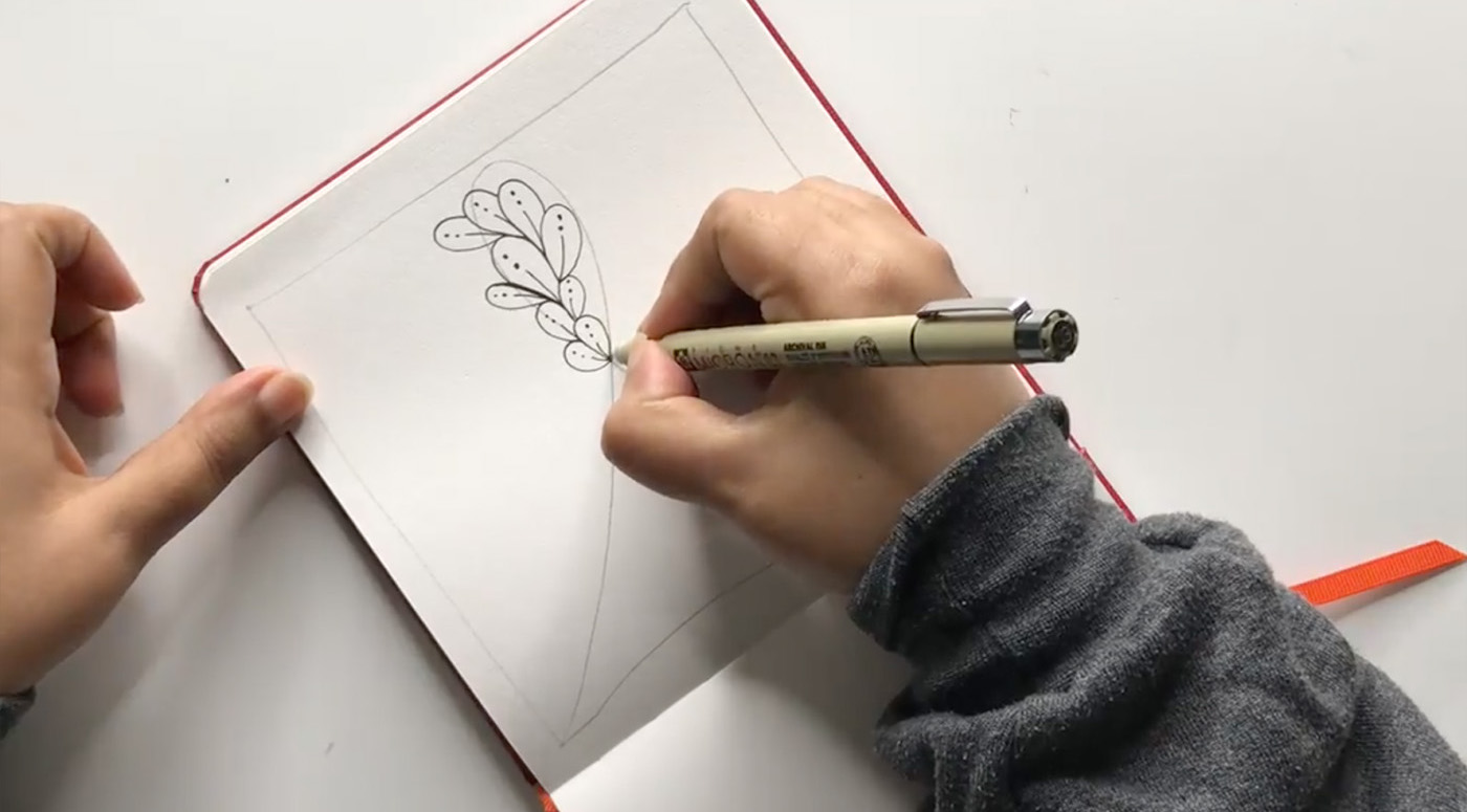 With only their two hands visible, someone draws an oval-shaped pattern on a notebook. 