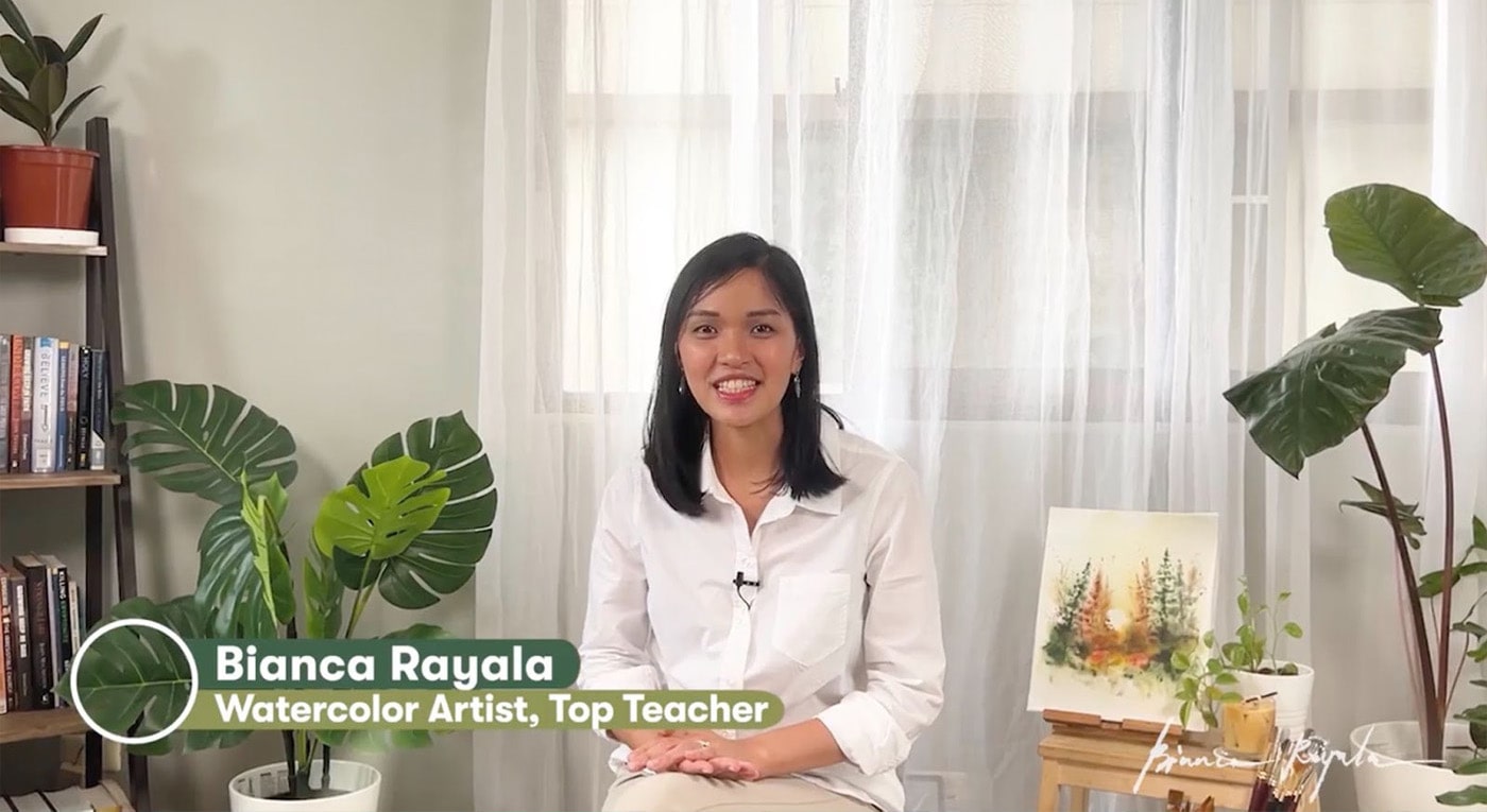 Teacher Bianca Rayala sits in front of a window covered by white curtains. One of her watercolor paintings, featuring a forest, is on an easel next to her, and some plants and a bookshelf sit nearby.