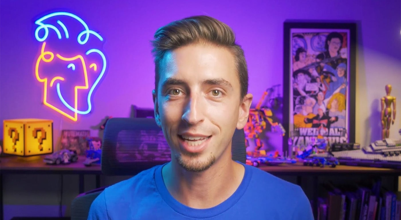 Teacher Jake Bartlett sits in his office. Behind him is a bookshelf, a neon sign in the shape of his face, and a variety of decorative items including posters, figurines, and a model of the question mark cube from Nintendo’s Mario games.