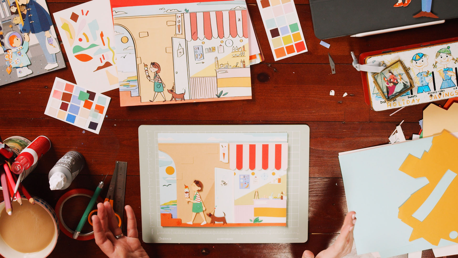 A papercraft vignette shows a little girl holding an ice cream cone after leaving the ice cream shop with her dog. The artwork is surrounded by the tools and materials needed to create it such as scraps of paper, a mug of colored pencils, scissors, tape, and the inspiration image.