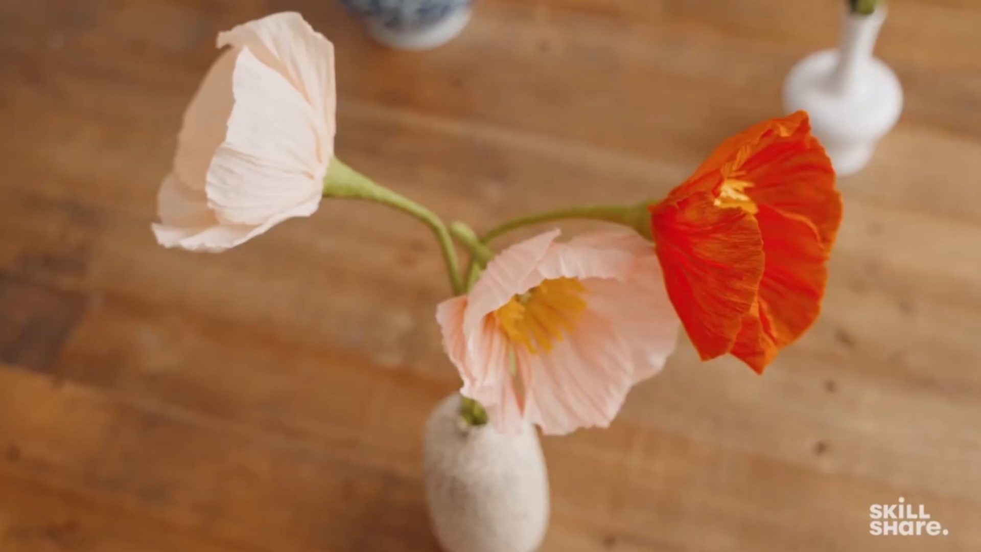Vase holding three red and pink paper poppies on a wooden table.