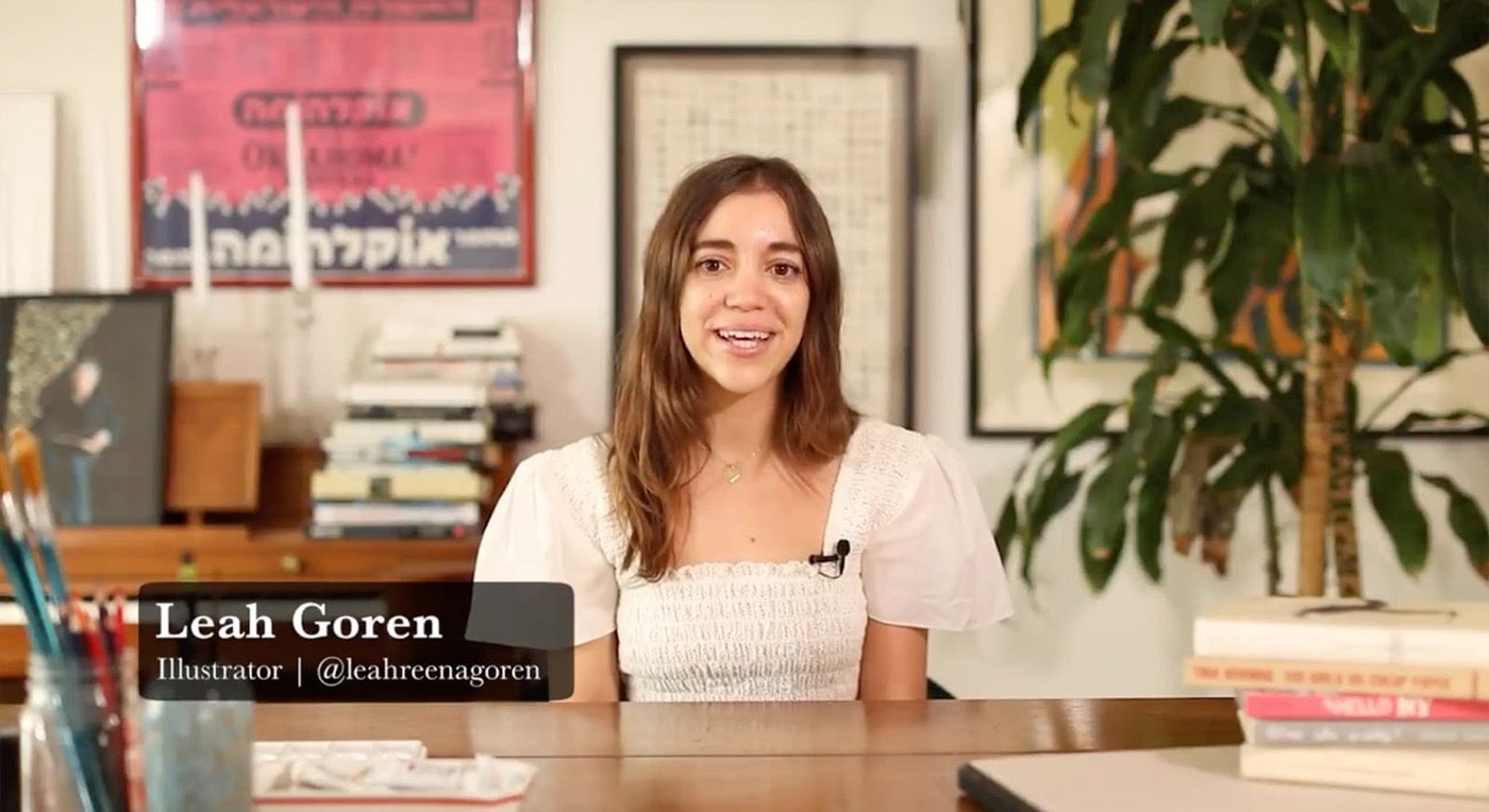 Teacher Leah Gorden sits at her desk. Books and jars of paint brushes sit in the front of the frame, and behind her we can see an upright piano, a few plants, and some photographs, posters and books.